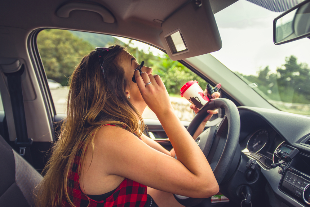 Young reckless female is sitting in her car while is doing her makeup on the car mirror
